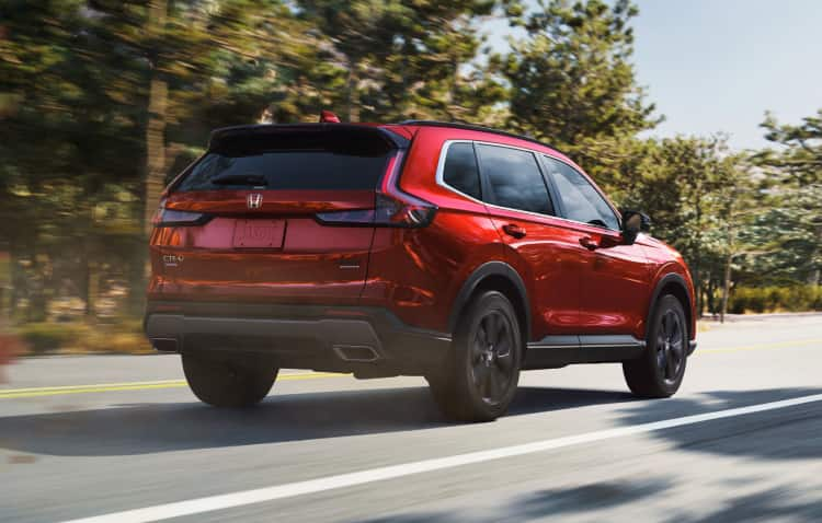 Vista trasera desde el lado del pasajero de la CR-V Sport 2025 con tracción integral en Radiant Red Metallic conduciendo por una carretera bordeada de árboles, levantando polvo y hojas